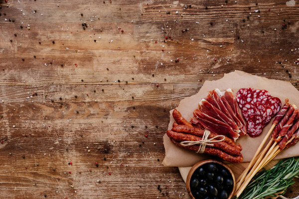 top view of round cutting board with delicious prosciutto, salami, smoked sausages, olives and herbs on wooden vintage table with scattered peppercorns