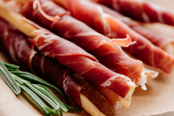 close up view of rosemary and delicious sliced prosciutto wrapped around breadsticks  on brown wrapping paper