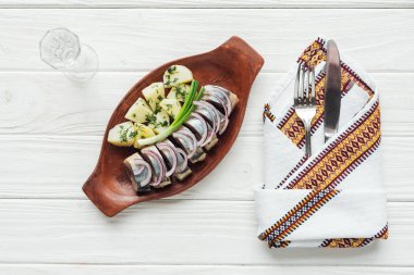 marinated herring, potatoes and onions in earthenware plate with glass of vodka, cutlery and embroidered towel on white wooden background clipart
