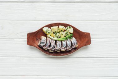 top view of marinated herring with potatoes and onions in earthenware plate on white wooden background clipart