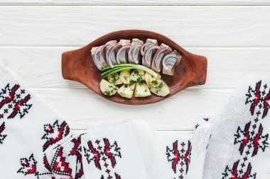 delicious marinated herring with potatoes and onions in earthenware plate with embroidered towel on white wooden background clipart