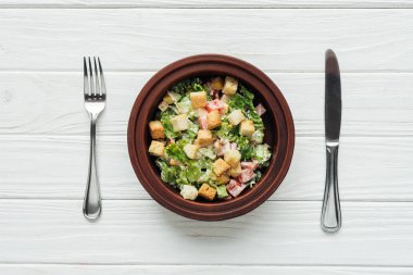 Beyaz ahşap zemin üzerinde lezzetli geleneksel Sezar salatası kase ve çatal bıçak takımı croutons üstten görünüm