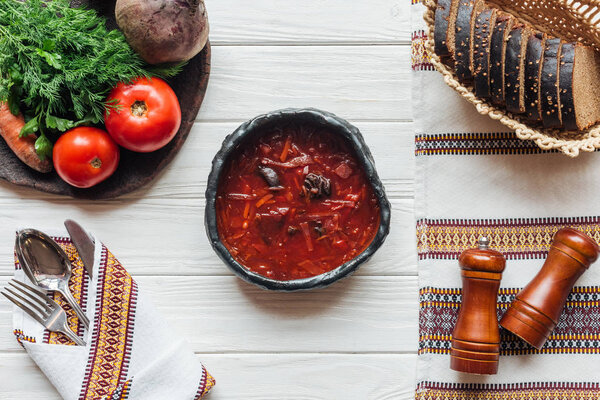 top view of traditional beetroot soup with cutlery, ingredients and rye bread on white wooden background