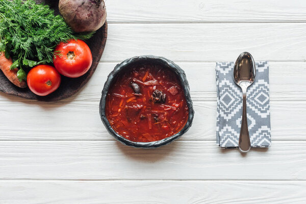 top view of traditional beetroot soup with spoon and ingredients on white wooden background