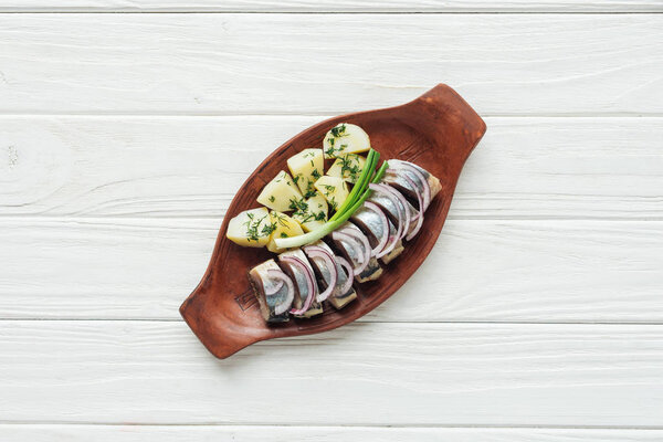 top view of marinated herring with potatoes and onions in earthenware plate on white wooden background
