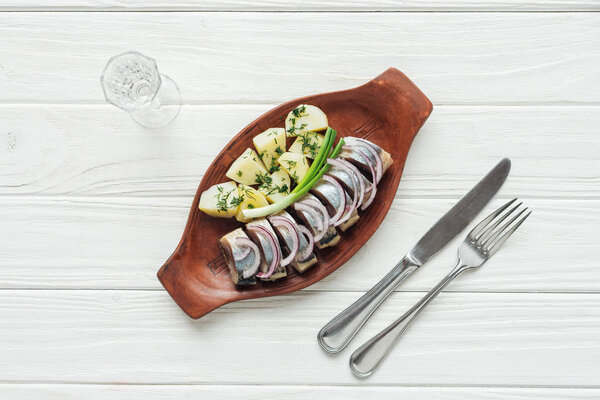 top view of marinated herring, potatoes and onions in earthenware plate with glass of vodka and cutlery on white wooden background