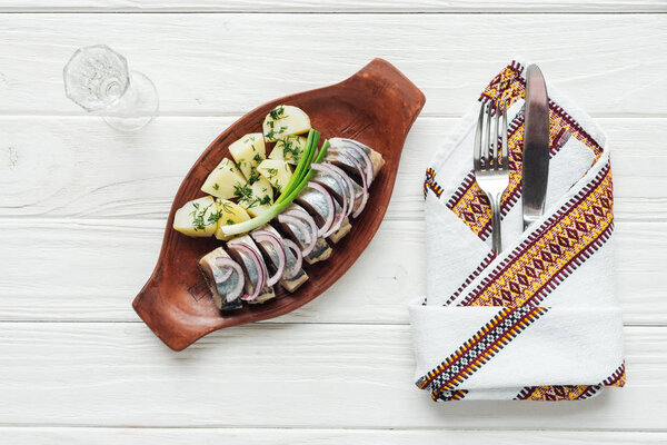 marinated herring, potatoes and onions in earthenware plate with glass of vodka, cutlery and embroidered towel on white wooden background