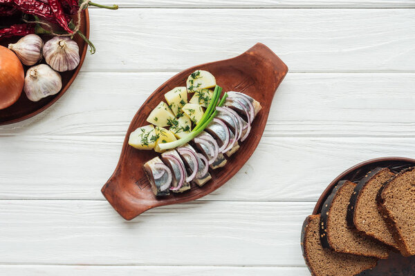 top view of marinated herring with potatoes and onions in earthenware plate with rye bread on white wooden background