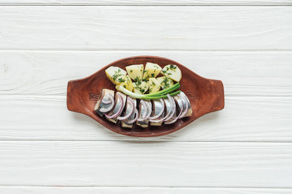 top view of marinated herring with potatoes and onions in earthenware plate on white wooden background