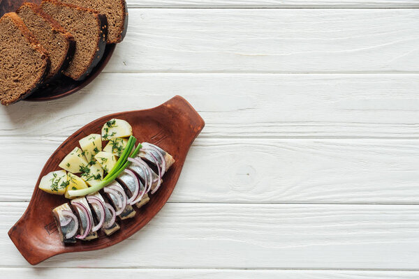 marinated herring with potatoes and onions in earthenware plate with rye bread and copy space on white wooden background