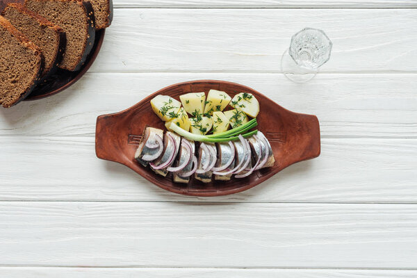 marinated herring, potatoes and onions in earthenware plate with glass of vodka and rye bread on white wooden background