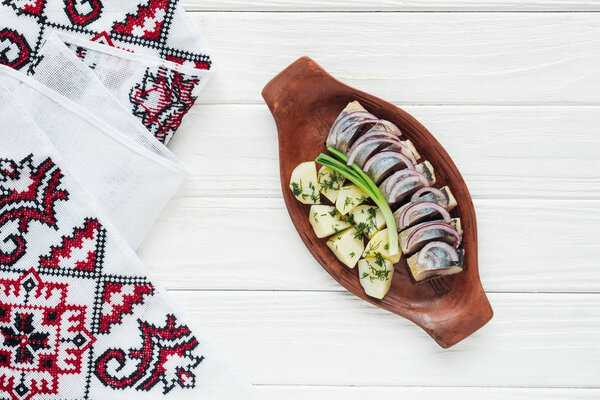 top view of marinated herring with potatoes and onions in earthenware plate with embroidered towel on white wooden background
