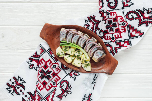 traditional marinated herring with potatoes and onions in earthenware plate with embroidered towel on white wooden background