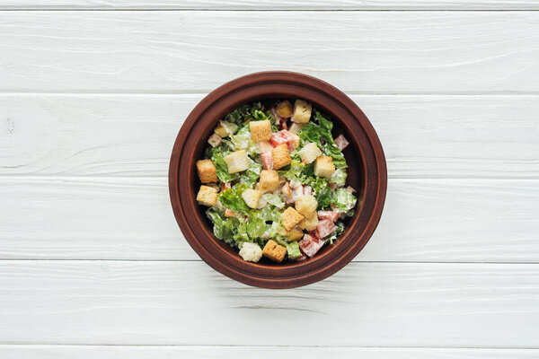 top view of tasty caesar salad with croutons in bowl on white wooden background