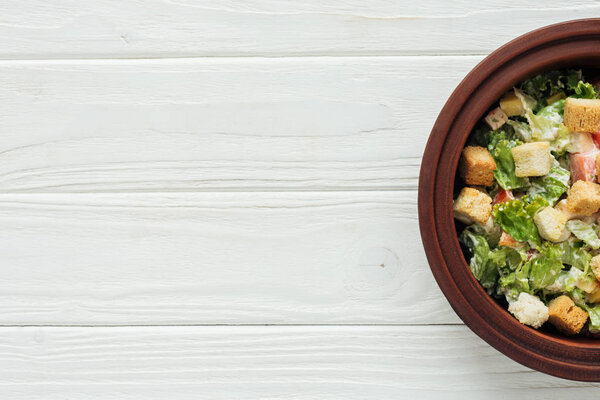 top view of traditional caesar salad with croutons in bowl on white wooden background with copy space