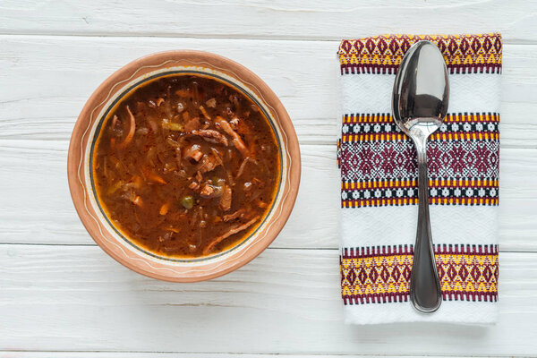 top view of delicious traditional mixed meat soup with spoon and embroidered towel on white wooden background