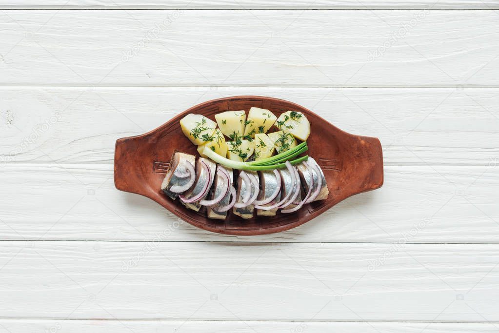top view of marinated herring with potatoes and onions in earthenware plate on white wooden background