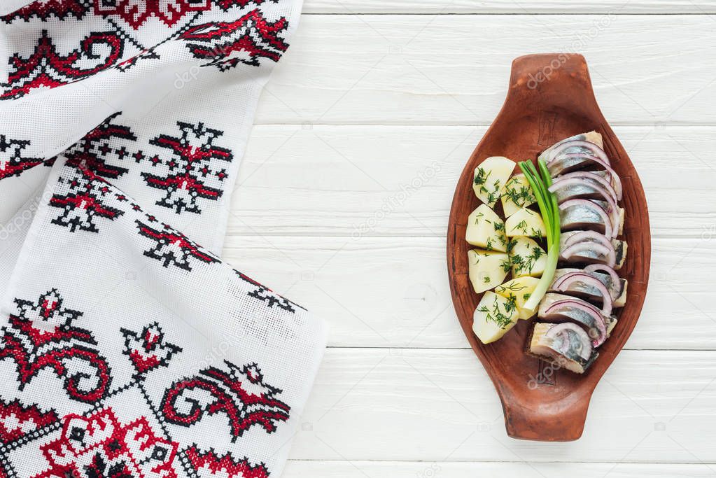 top view of marinated herring with potatoes and onions in earthenware plate with embroidered towel on white wooden background