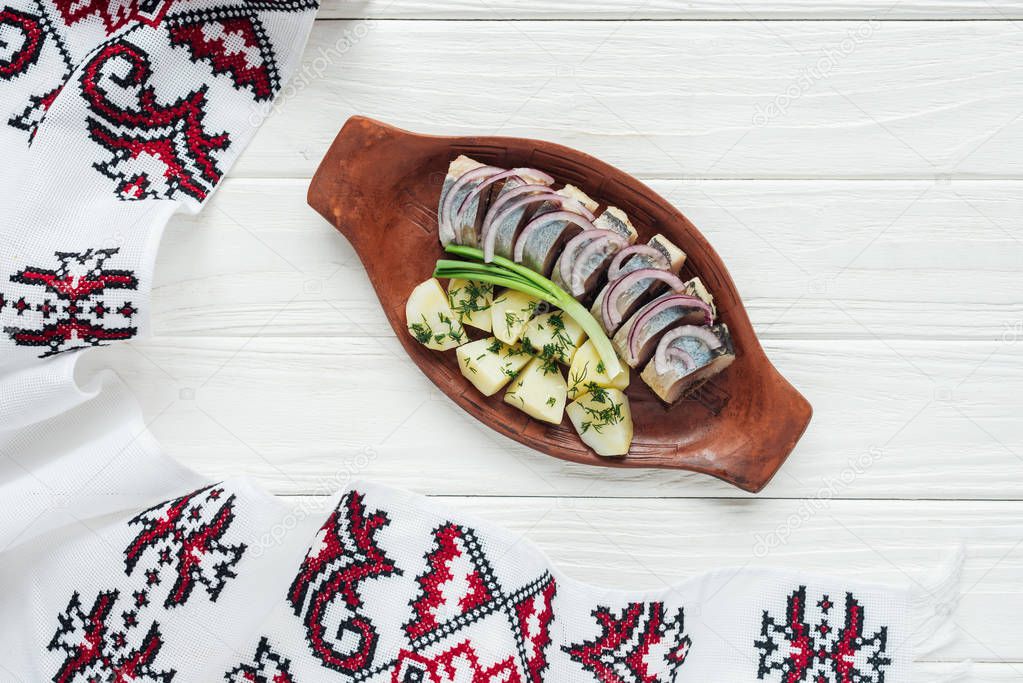 traditional marinated herring with potatoes and onions in earthenware plate with embroidered towel on white wooden background