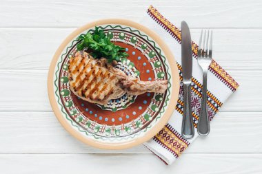 delicious rib eye meat steak on plate with parsley, cutlery and embroidered towel on white wooden background clipart