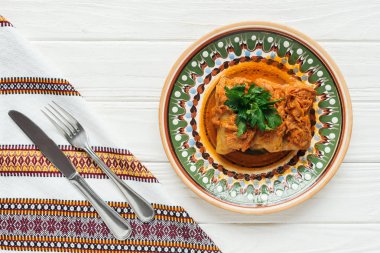 top view of delicious stuffed cabbage rolls with parsley, cutlery and embroidered towel on white wooden background clipart