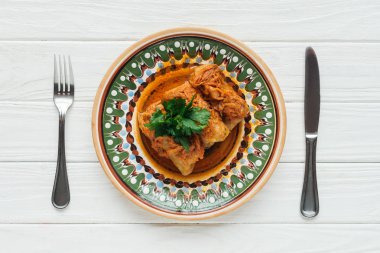 top view of tasty stuffed cabbage rolls with parsley on plate, cutlery and white wooden background clipart