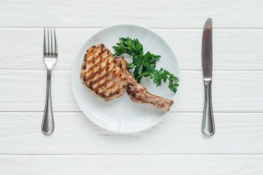 top view of tasty rib eye meat steak on plate with parsley and cutlery on white wooden background clipart