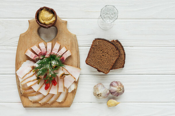 top view of traditional sliced smoked lard on cutting board with mustard, glass of vodka and rye bread on white wooden background