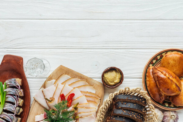 top view of traditional sliced smoked lard on cutting board, rye bread, herring and mini pies on white wooden background