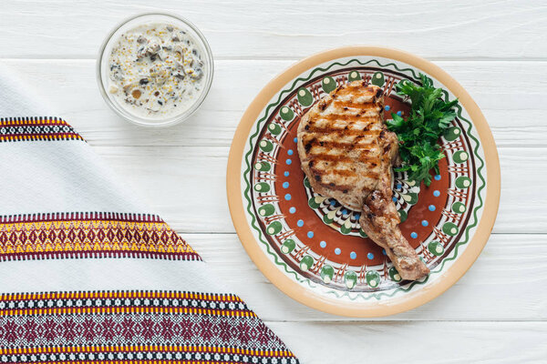 tasty rib eye meat steak on plate with parsley, sauce and embroidered towel on white wooden background