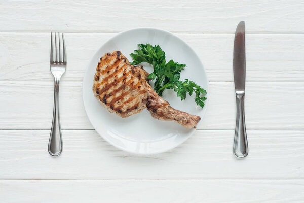 top view of tasty rib eye meat steak on plate with parsley and cutlery on white wooden background