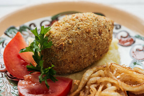 close up of chicken kiev with mashed potatoes, parsley, tomatoes and fried onions on plate with ornament