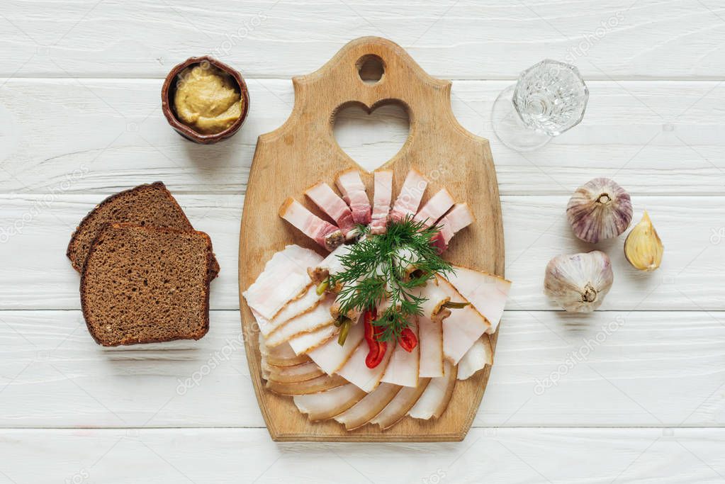 traditional sliced smoked lard on cutting board with mustard, glass of vodka and rye bread on white wooden background
