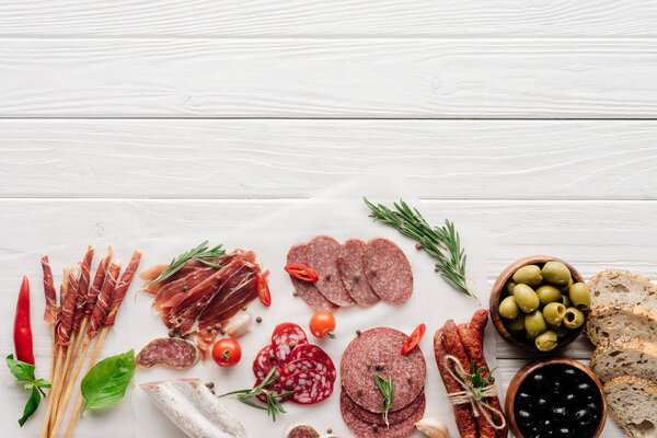 top view of arrangement of various meat snacks and olives on white wooden backdrop
