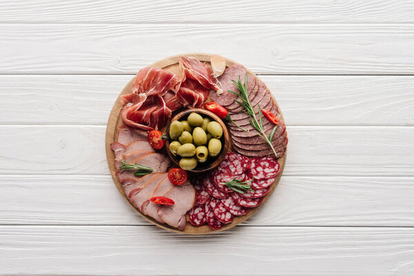 top view of arrangement of various meat snacks and olives on white wooden backdrop