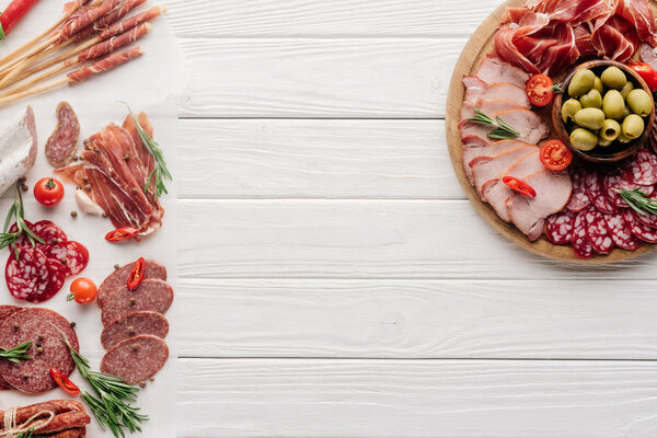 top view of arrangement of various meat snacks and olives on white wooden backdrop