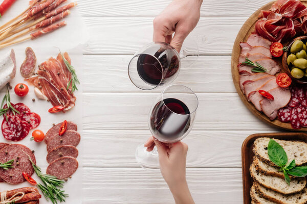 partial view of couple clinking glasses with red wine at white tabletop with meat appetizers