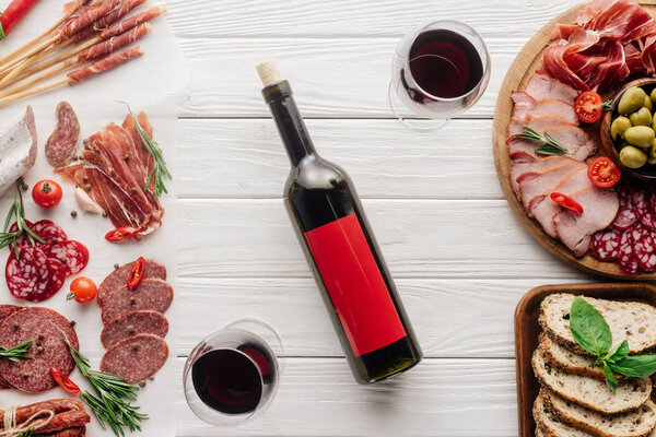 food composition with bottle and glasses of red wine, pieces of bread, olives and meat snacks on white wooden tabletop