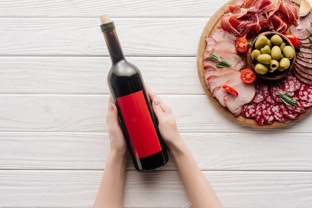 partial view of woman holding bottle of red wine on white tabletop with meat appetizers