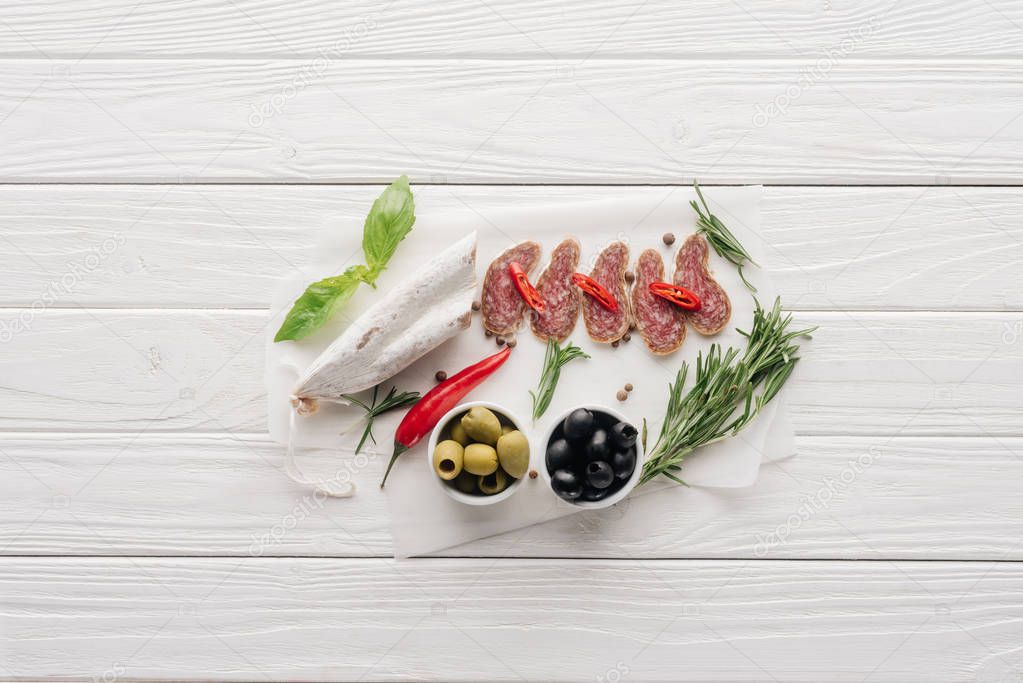 top view of meat appetizers with olives, rosemary and basil leaves on white wooden tabletop