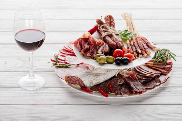 close up view of olives, glass of red wine and assorted meat snacks on white wooden tabletop