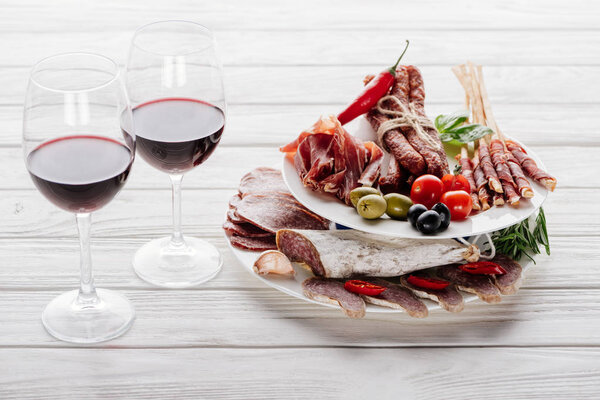 close up view of tasty meat appetizers and glasses of red wine on white wooden surface