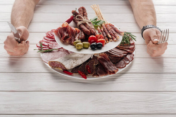 partial view of man with cutlery at white wooden tabletop with various meat snacks