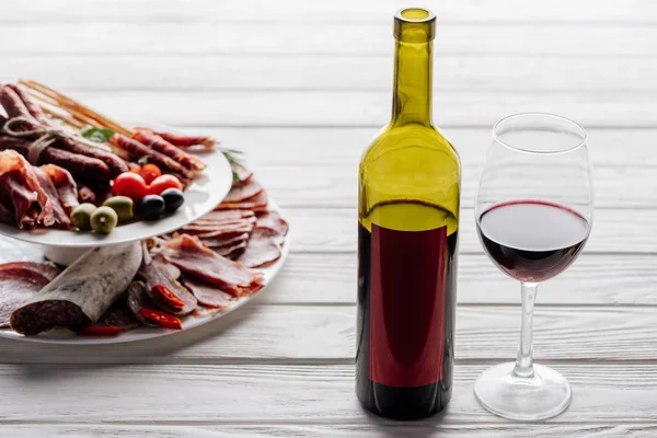 靠近葡萄酒瓶和一杯红酒和各种肉类开胃菜与橄榄在白色木制桌面上的特写视图 — 图库照片