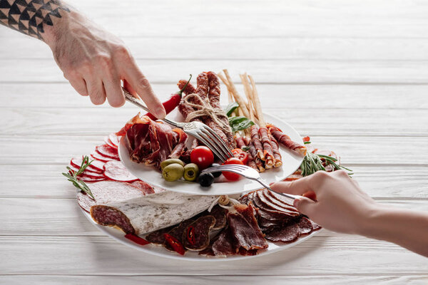 cropped shot of couple trying meat appetizers at white wooden background