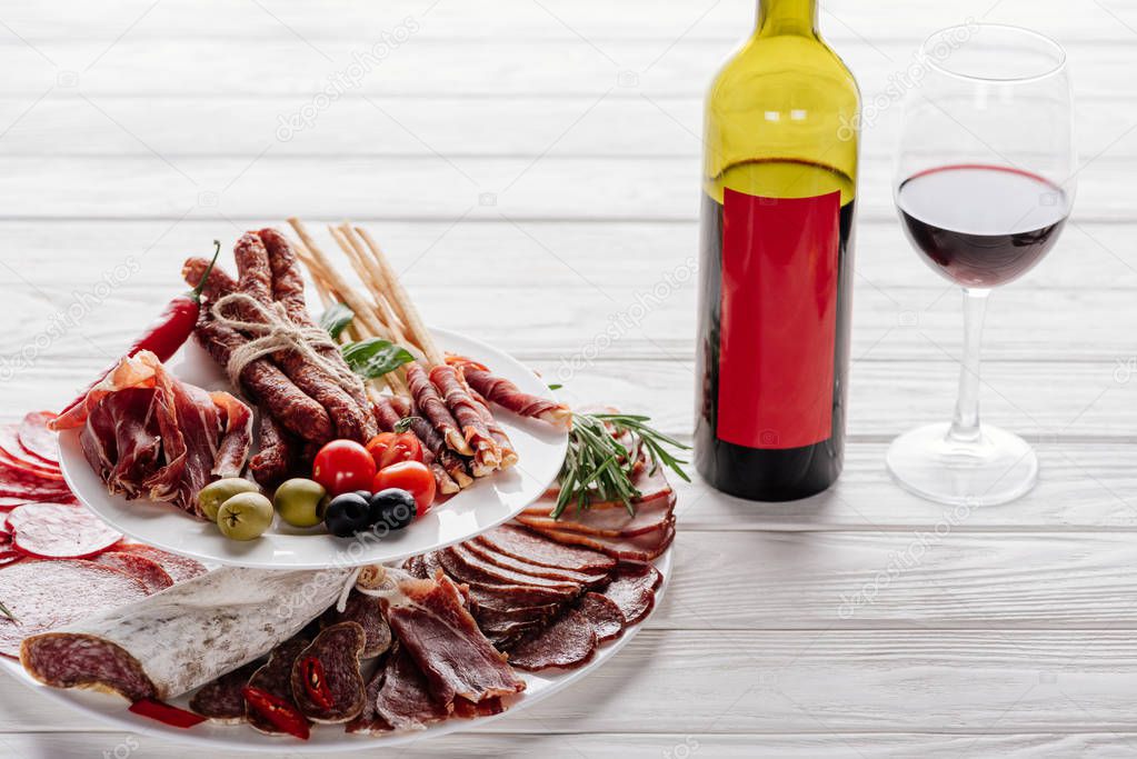 close up view of bottle and glass of red wine and various meat appetisers with olives on white wooden tabletop