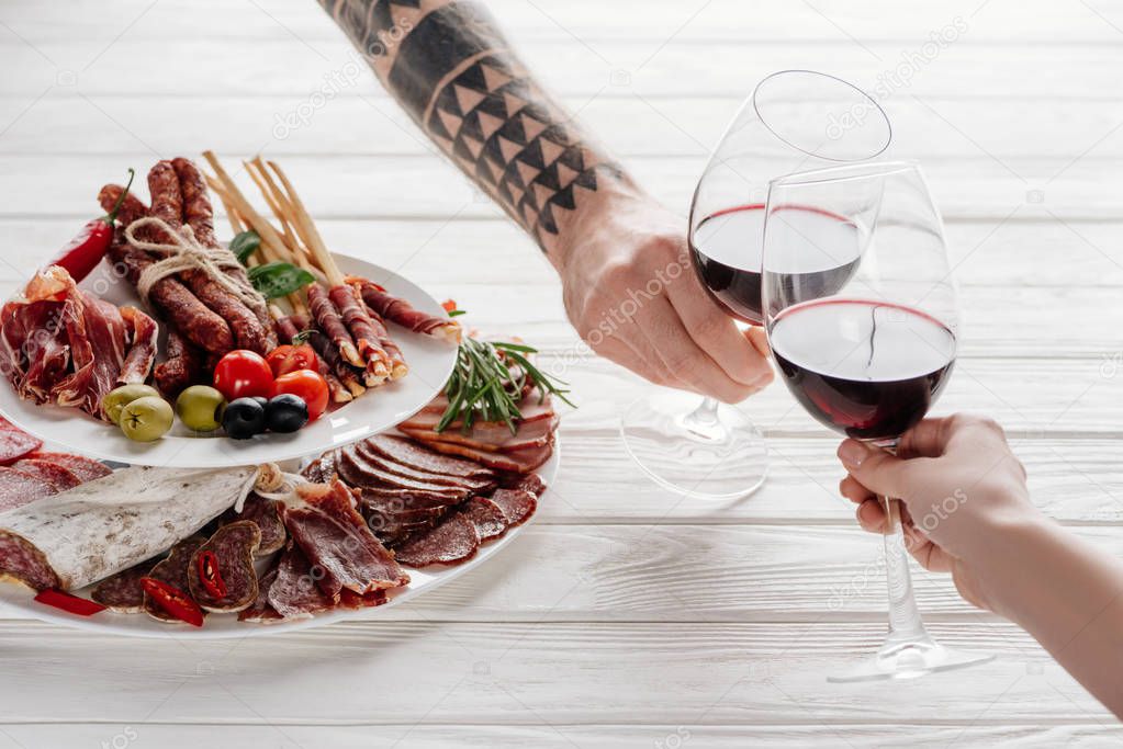 partial view of couple clinking glasses with red wine at white tabletop with meat appetizers