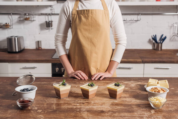 partial view of woman in apron preparing triple chocolate mousse in glasses