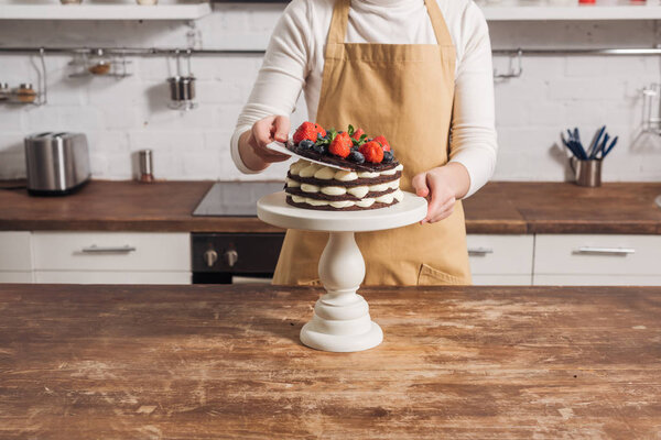 cropped shot of woman in apron preparing delicious cake with fruits  