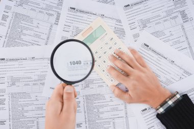 cropped view of man holding magnifying glass and using calculator with tax forms on background  clipart